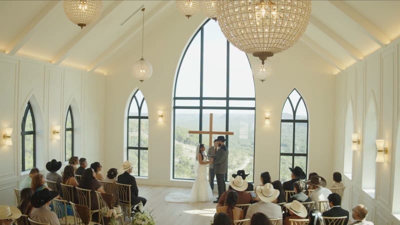 A couple saying their vows at the altar of the chapel, framed by the arched window and Hill Country view, with guests seated on both sides