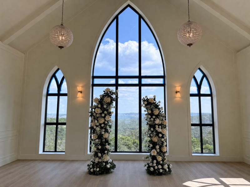 Two lush white floral and greenery pillars framing the chapel's arched window as a ceremony backdrop