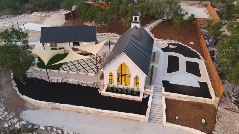 Aerial view of The Chapel at Wimberley beside its gathering courtyard with cream shade sails, a checkerboard lawn, and the bell house