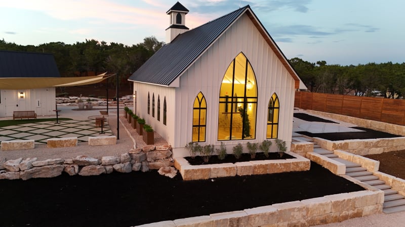 Side of The Chapel at Wimberley at dusk with its tall gothic arched window glowing warm, limestone terraces and the gathering courtyard's shade sails beside it
