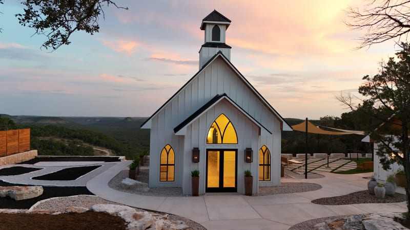 Front of The Chapel at Wimberley at sunset, a white board-and-batten wedding chapel with a steeple and glowing gothic arched windows, the Texas Hill Country rolling away behind it