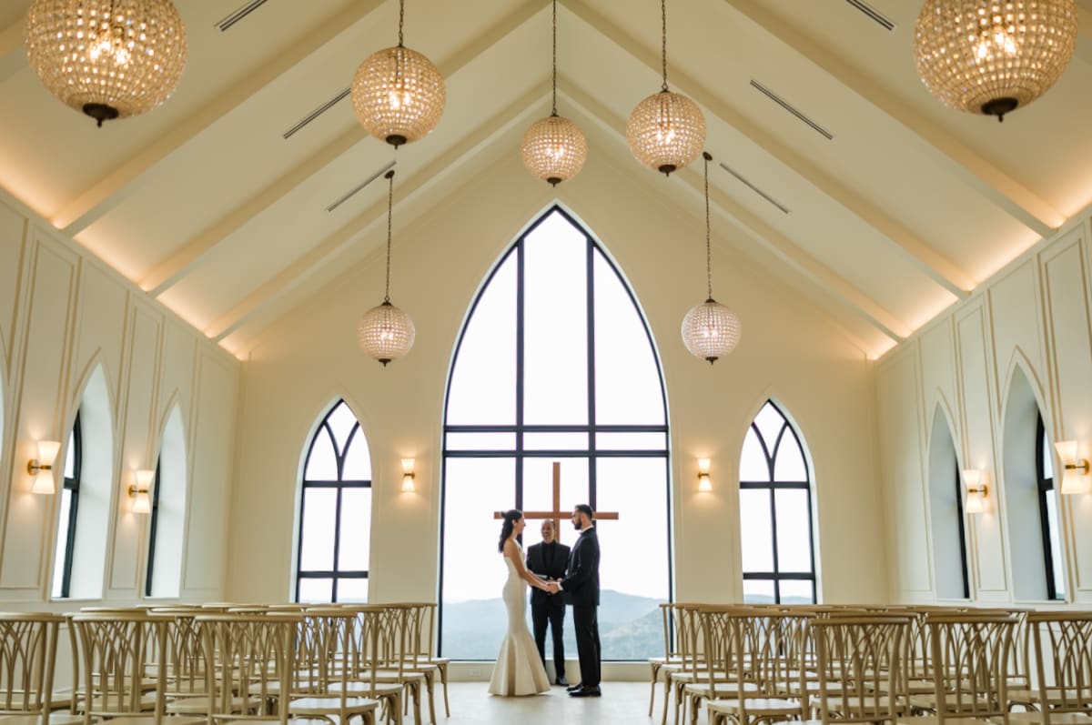 A couple exchanges vows at the altar of The Chapel at Wimberley beneath crystal chandeliers, framed by the great arched window and the Hill Country beyond