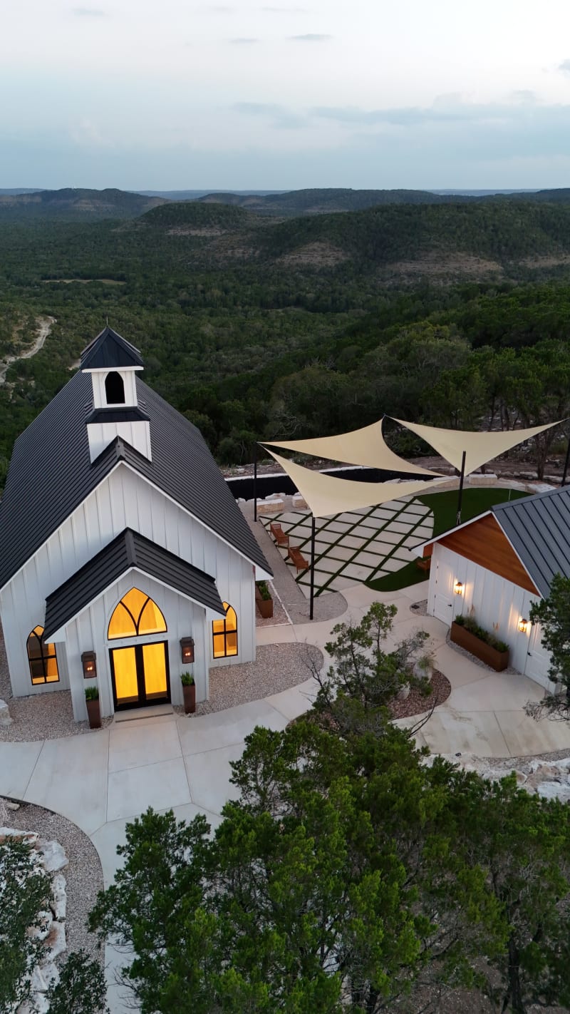 Aerial view of The Chapel at Wimberley at dusk, its windows glowing, with the gathering courtyard and shade sails beside it and Hill Country ridges rolling to the horizon